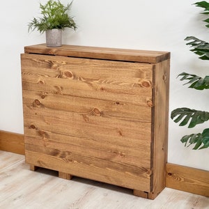 May include: A wooden folding table with a natural brown finish. The table is closed and standing upright against a white wall. The table has a simple design with a rectangular top and a base with four legs.