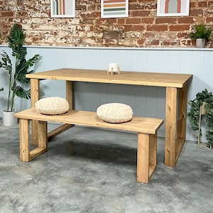 May include: A wooden dining table and bench set with two fluffy cushions. The table and bench are made of natural wood with a light brown finish. The table has a rectangular top and four legs. The bench has a rectangular top and two legs on each side. The cushions are white and fluffy. The set is in front of a brick wall with three abstract art prints.