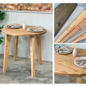 May include: A round wooden table with four legs. The table is made of natural wood and has a light brown finish. The table is set with two place settings, each with a white plate, a beige napkin, and a small vase with a single flower.