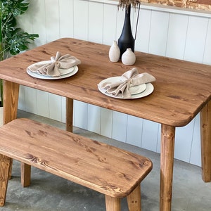 May include: A wooden dining table with a brown finish and a matching bench. The table has a rounded edge and is set with two white plates and napkins. A vase with dried flowers and two small ceramic vases are on the table.