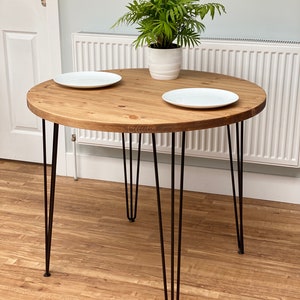 May include: A round wooden table with a natural wood finish and black hairpin legs. Two white plates are placed on the table, and a potted plant sits in the centre.