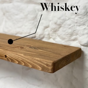 May include: A rustic wooden shelf with a natural brown stain. The shelf is mounted on a white brick wall. The word "Whiskey" is written in black text above the shelf.