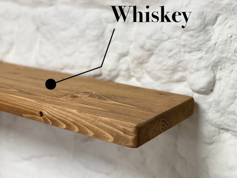 May include: A rustic wooden shelf with a natural brown stain. The shelf is mounted on a white brick wall. The word "Whiskey" is written in black text above the shelf.