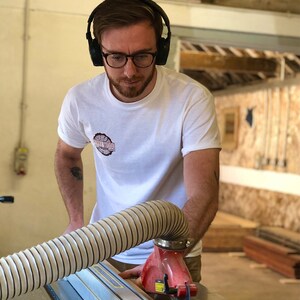 May include: A man wearing headphones and glasses is operating a table saw in a woodworking shop. He is wearing a white t-shirt with the text "Sawdust Wood" printed on it.