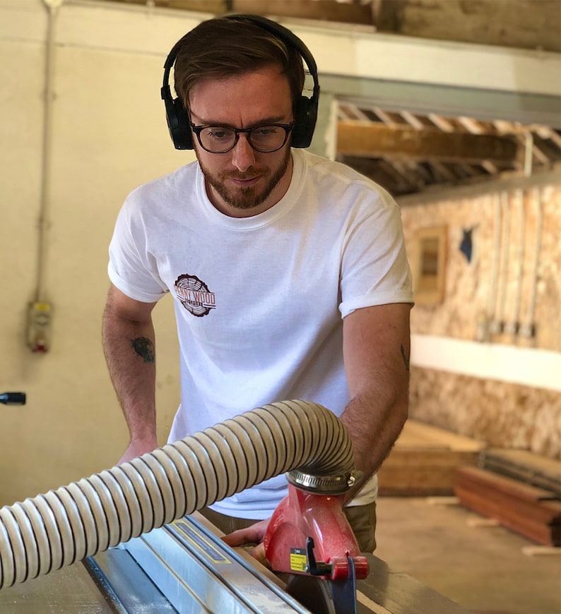 May include: A man wearing headphones and glasses is operating a table saw in a woodworking shop. He is wearing a white t-shirt with the text "Sawdust Wood" printed on it.