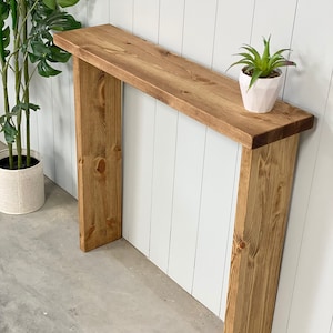 May include: A rustic wooden console table with a natural wood finish. The table has a simple design with two vertical supports and a rectangular top. A small potted plant sits on the table.