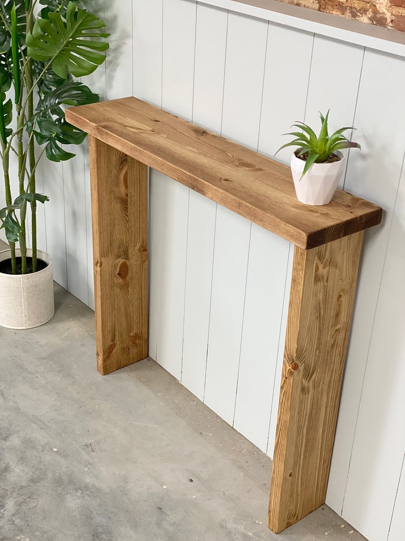 May include: A rustic wooden console table with a natural wood finish. The table has a simple design with two vertical supports and a rectangular top. A small potted plant sits on the table.
