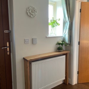 May include: A white radiator with a wooden shelf above it. The shelf is made of reclaimed wood and has a natural finish. The radiator is in a hallway with a white wall and a door to the left.