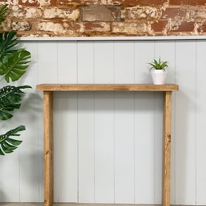 May include: A rustic wooden console table with a natural wood finish. The table has a simple design with a rectangular top and two vertical legs. A small potted plant sits on the table top.