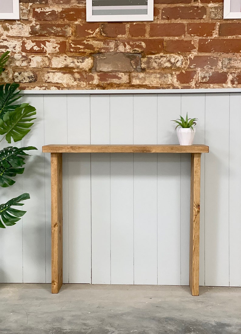 May include: A rustic wooden console table with a natural wood finish. The table has a simple design with a rectangular top and two vertical legs. A small potted plant sits on the table top.
