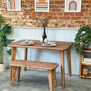 May include: A wooden dining table and bench set with a rustic brown finish. The table has a rectangular top and four legs. The bench has a rectangular seat and four legs. The set is placed in front of a brick wall with three framed abstract prints.