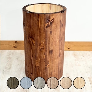May include: A tall, cylindrical wooden container with a dark brown stain. The container has a natural wood grain pattern and a light-coloured interior. The top edge is reinforced with metal hardware. The base of the container has a selection of colour swatches.