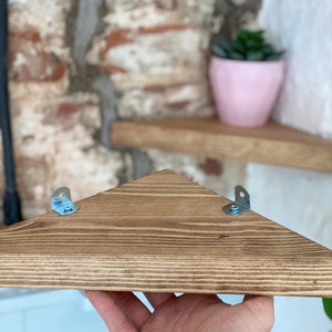 May include: A triangular wooden shelf with a dark brown stain. The shelf has two metal brackets on the underside for mounting.