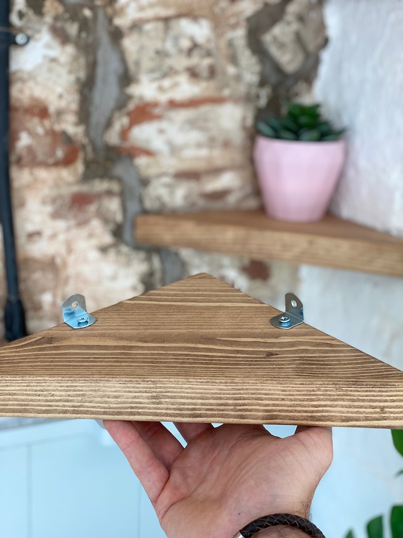 May include: A triangular wooden shelf with a dark brown stain. The shelf has two metal brackets on the underside for mounting.