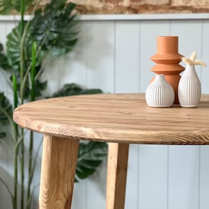 May include: A round wooden table with a light brown finish and three legs. The table is topped with three decorative vases, one orange and two white.