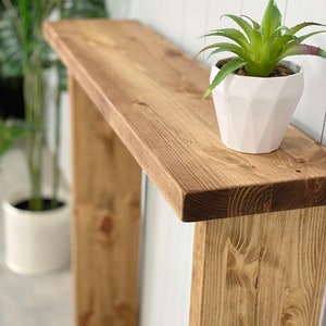 May include: A rustic wooden console table with a natural wood finish. The table has a single shelf and two legs. A white planter with a green succulent plant sits on the shelf.