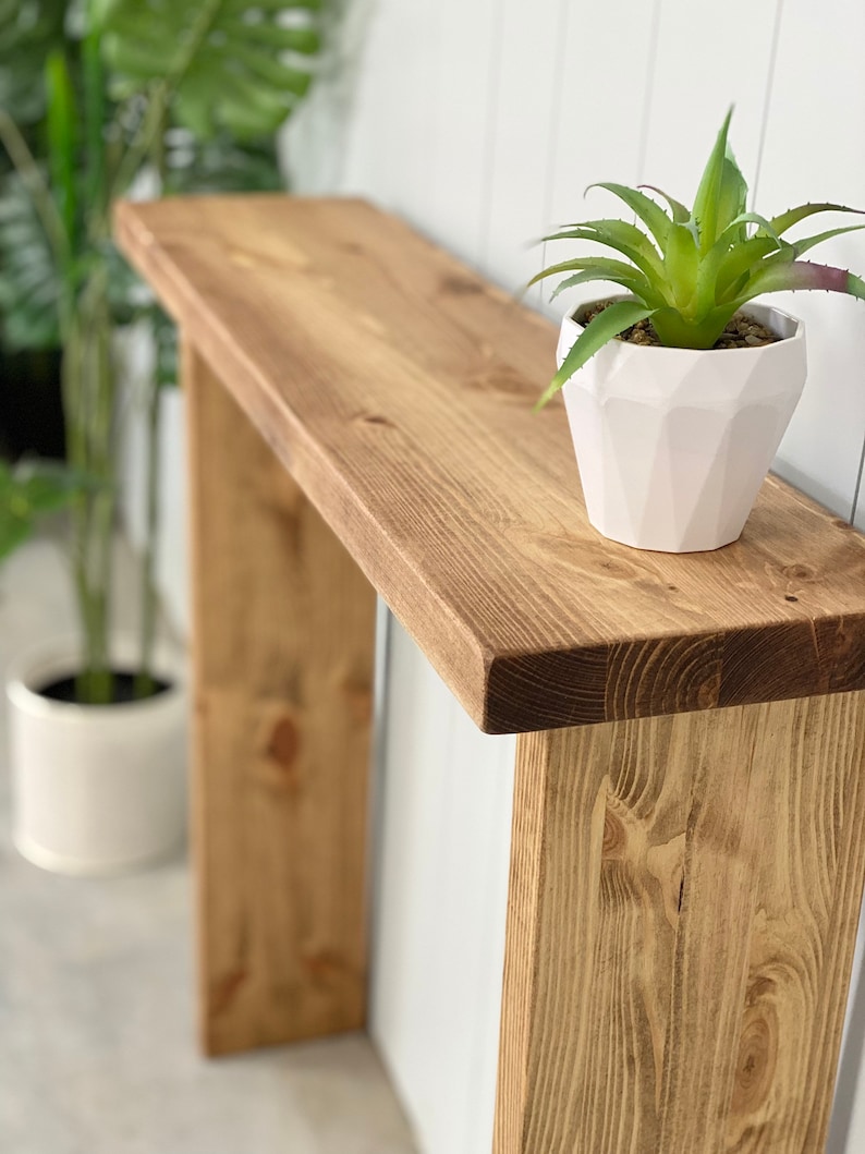 May include: A rustic wooden console table with a natural wood finish. The table has a single shelf and two legs. A white planter with a green succulent plant sits on the shelf.
