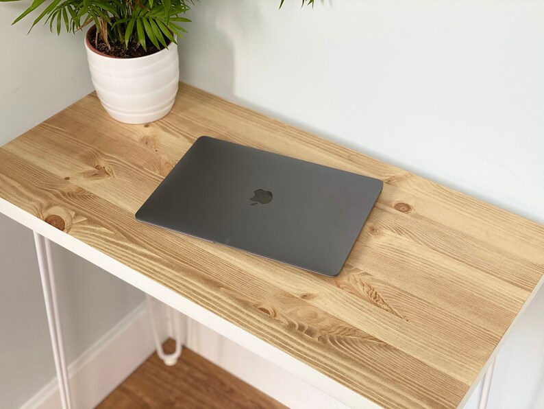 May include: A light wood desk with a white metal frame and a laptop computer on top. The desk is in a room with white walls and a potted plant.