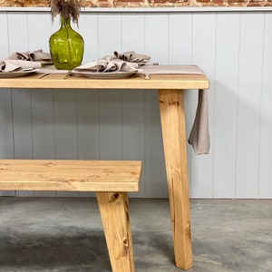 May include: A wooden dining table with a natural finish and a matching bench. The table has four legs and is set with a linen tablecloth and napkins. A green glass vase with dried flowers sits on the table.