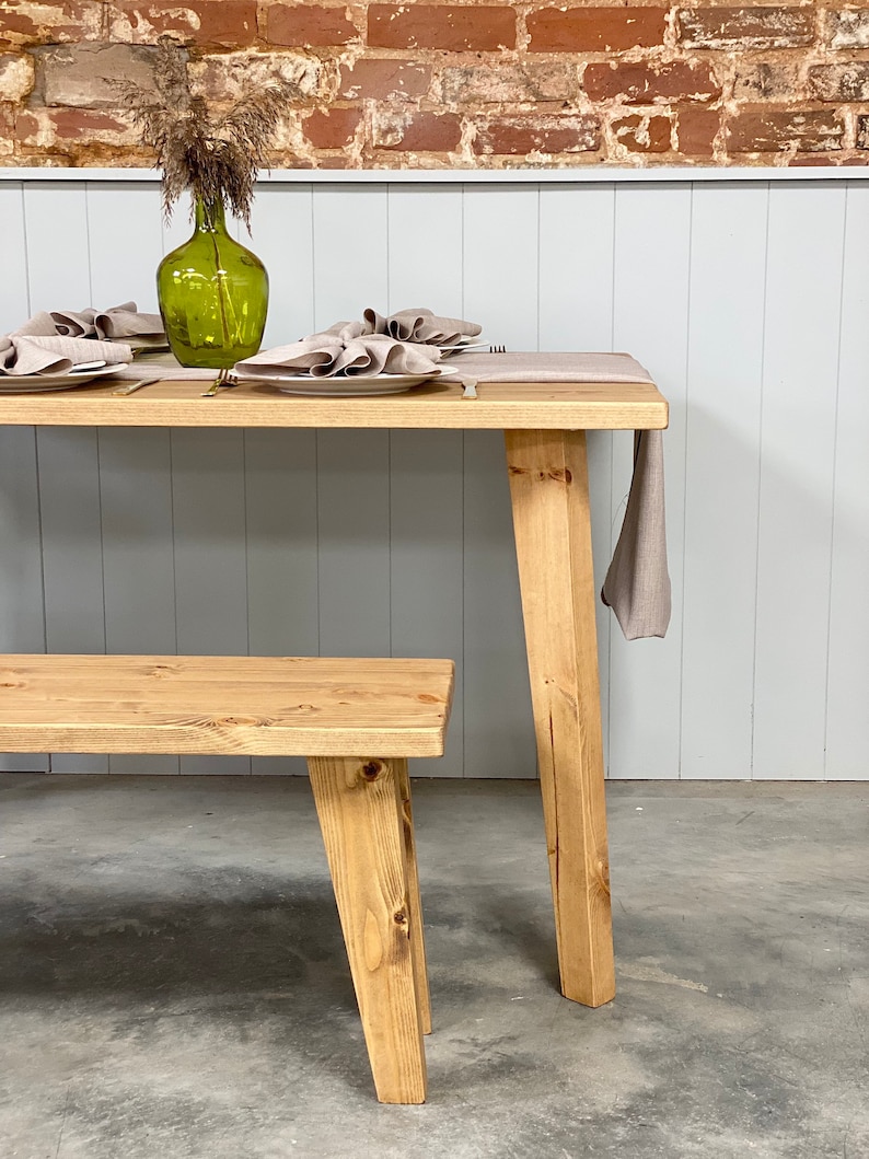 May include: A wooden dining table with a natural finish and a matching bench. The table has four legs and is set with a linen tablecloth and napkins. A green glass vase with dried flowers sits on the table.