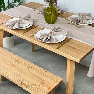 May include: A rustic wooden table with a light brown finish and a matching bench. The table is set with white plates, gold silverware, and linen napkins. A vase with dried flowers sits in the center of the table.
