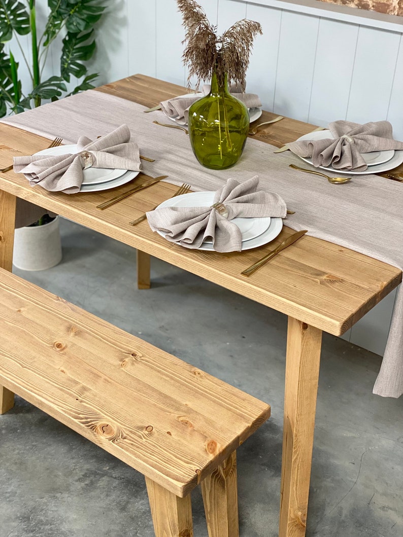 May include: A rustic wooden table with a light brown finish and a matching bench. The table is set with white plates, gold silverware, and linen napkins. A vase with dried flowers sits in the center of the table.