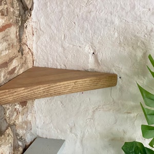 May include: A triangular wooden shelf mounted on a brick wall. The shelf is stained a light brown color and has a natural wood grain. The shelf is mounted in a corner of the wall.