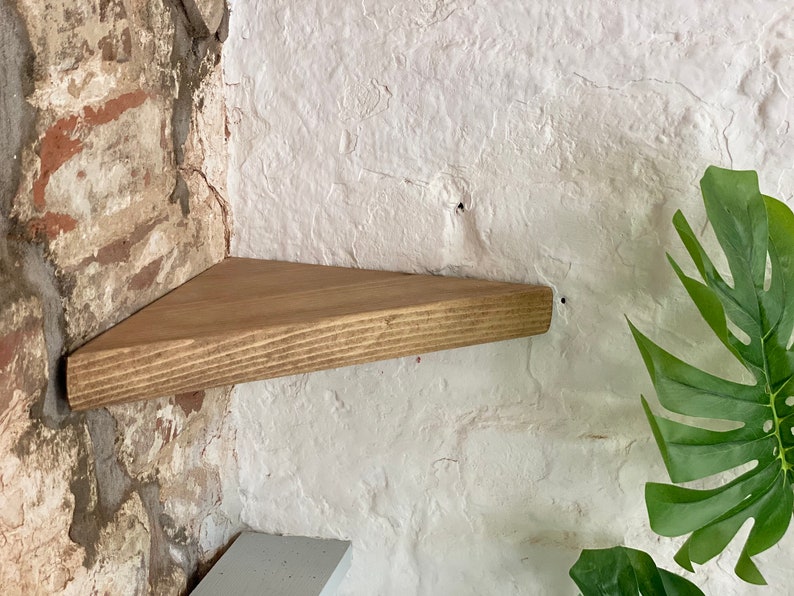 May include: A triangular wooden shelf mounted on a brick wall. The shelf is stained a light brown color and has a natural wood grain. The shelf is mounted in a corner of the wall.