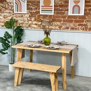 May include: A wooden dining table with a natural finish and a matching bench. The table has a light brown finish and is set with three place settings. The bench is also made of wood and has a natural finish. There are three framed prints on the wall behind the table, each with a different abstract design.