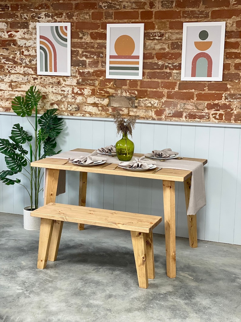 May include: A wooden dining table with a natural finish and a matching bench. The table has a light brown finish and is set with three place settings. The bench is also made of wood and has a natural finish. There are three framed prints on the wall behind the table, each with a different abstract design.