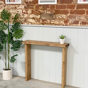 May include: A rustic wooden console table with a natural wood finish. The table has a rectangular top and two legs. The table is standing in front of a white wall with a brick wall behind it.