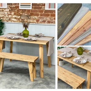 May include: A rustic wooden table and bench set with a light brown finish. The table is set with a light gray linen tablecloth and napkins, a green glass vase with greenery, and gold flatware. The bench is made of the same wood as the table and has a simple design.