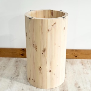 May include: A cylindrical wooden side table or stool. The light-coloured wood has visible knots and vertical grain patterns. The top edge has small metal fasteners. The piece is approximately 61 cm tall and 41 cm in diameter.