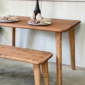 May include: A wooden dining table with a natural finish and four legs. A matching wooden bench is in the foreground. The table is set with two plates and napkins.