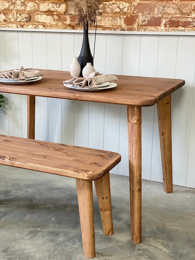 May include: A wooden dining table with a natural finish and four legs. A matching wooden bench is in the foreground. The table is set with two plates and napkins.