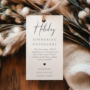 Könnte beinhalten: Ein cremefarbenes Etikett mit den Worten "Holiday Simmering Potpourri" in eleganter Schrift. Das Etikett enthält Anweisungen und ist mit Bindfaden gebunden, umgeben von getrockneten Blumen und Blättern.
