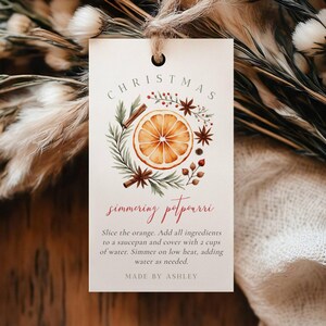 May include: A rectangular tag with the words "CHRISTMAS simmering potpourri" in a decorative font. The tag features a watercolor illustration of an orange slice, cinnamon sticks, and star anise. Instructions are printed below the illustration.