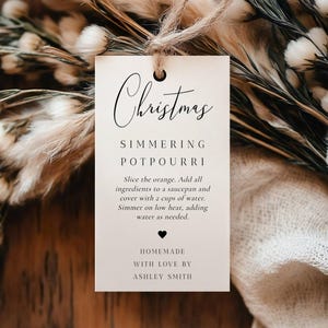 Puede incluir: Una etiqueta color crema con la palabra "Christmas" en escritura elegante, que detalla una receta de popurrí a fuego lento. La etiqueta está atada con hilo y rodeada de flores y follaje secos.