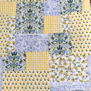 May include: A patchwork quilt featuring various floral and gingham patterns in shades of yellow, blue, and white. The quilt has a ruffled edge and is composed of multiple fabric squares with different designs.