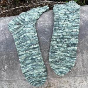 May include: A pair of hand-knitted socks in shades of blue, green, and grey. The socks feature a cable knit pattern and are lying on a weathered, grey surface. The socks are a cosy, handmade item.