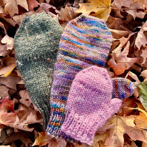May include: Three hand-knitted mittens in shades of green, purple, and pink rest on a bed of fallen autumn leaves. The mittens display various colour patterns and textures, showcasing the craft of knitting. The scene evokes warmth and the season of autumn.