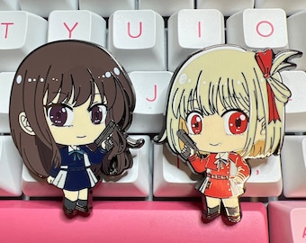 May include: Two enamel pins featuring anime-style female characters with guns. The character on the left has brown hair and is wearing a blue and white uniform. The character on the right has blonde hair and is wearing a red dress.