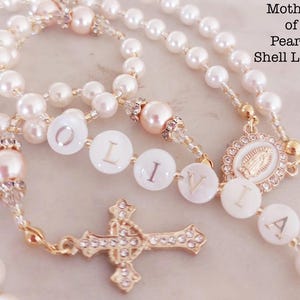 May include: A pearl rosary with the name "LIVIA" spelled out in white shell letters. The rosary features white and pink pearls, gold accents, and a decorative cross. The text "Mother of Pearls Shell Letter" is visible.