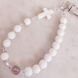 May include: White beaded rosary keychain with a silver clasp and a small cross pendant. The keychain features white beads, clear crystal accents and a silver medal. The beads are approximately 0.8 cm in diameter.