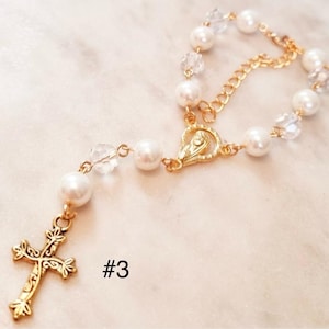 May include: A white pearl and crystal rosary bracelet with a gold chain and a gold cross charm. The bracelet has a gold clasp with a small image of the Virgin Mary. The text #3 is visible in the image.