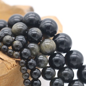 May include: A close-up of a pile of black and gold gemstone beads. The beads are round and have a smooth, polished finish. They are arranged in a random pattern, with some beads overlapping others.