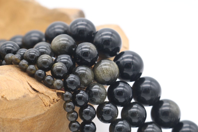 May include: A close-up of a pile of black and gold gemstone beads. The beads are round and have a smooth, polished finish. They are arranged in a random pattern, with some beads overlapping others.