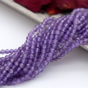 Könnte beinhalten: Eine Strähne aus violetten Amethystperlen, facettiert und rund, mit leicht unregelmäßiger Form. Die Perlen sind auf einem durchsichtigen Faden aufgereiht.