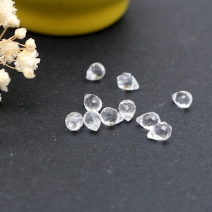 Könnte beinhalten: Eine Ansammlung kleiner, klarer, facettierter Edelsteine in Tropfenform. Diese transparenten Steine weisen mehrere geschliffene Oberflächen auf, die das Licht reflektieren, und sind in einer losen Gruppe neben einem Zweig weißer Trockenblumen angeordnet.