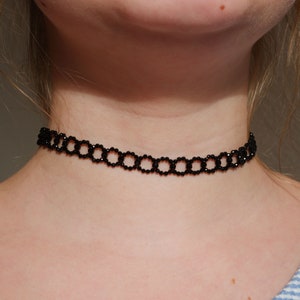 Könnte beinhalten: Eine schwarze Choker-Kette aus kleinen, runden Perlen. Die Kette wird von einer Person getragen, aber das Gesicht der Person ist nicht sichtbar.